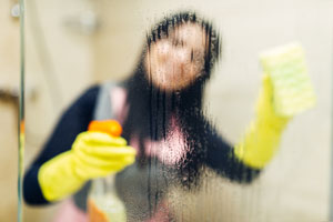 Image of woman's cleaning the shower with kitchen gloves. Article covers why the stress of living through the pandemic is causing weak, peeling and brittle fingernails. Nailcarehq.com