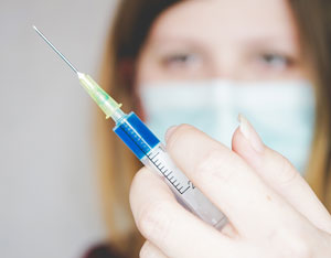 Image of woman wearing a paper disposable mask worn during the Covid quarantine pandemic and vaccine syringe. Article covers why the stress of living through the pandemic is causing weak, peeling and brittle fingernails. Nailcarehq.com